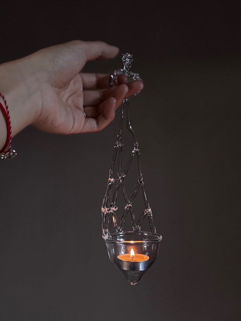 A hand holds the Suspended Candle Holder from the HolyEgg Series, crafted from clear wire and glass with a lit tealight inside, set against a plain dark background.